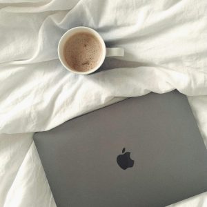 A cozy morning featuring a coffee cup and laptop on soft white bedding, viewed from above.
