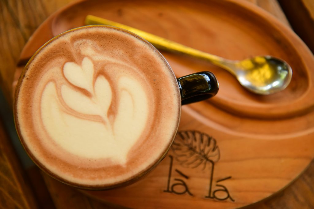 Intricate latte art coffee on a wooden tray with a spoon, in a cozy cafe setting.